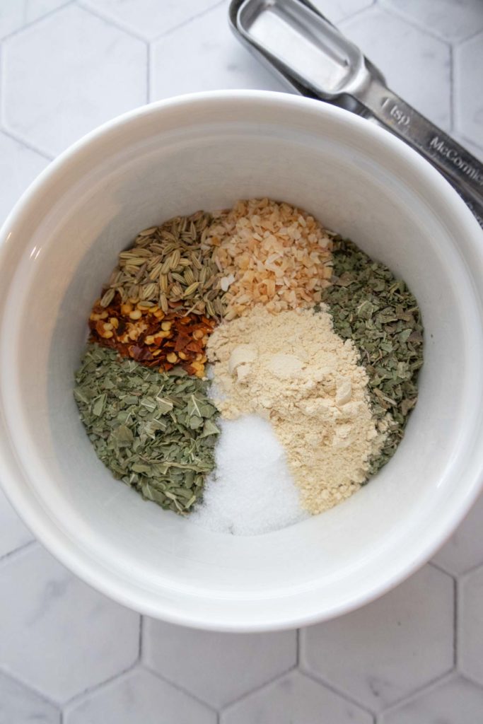 A white bowl containing a mix of dried herbs and spices, including fennel seeds, red pepper flakes, minced onion, oregano, garlic powder, and salt, with a measuring spoon beside it.