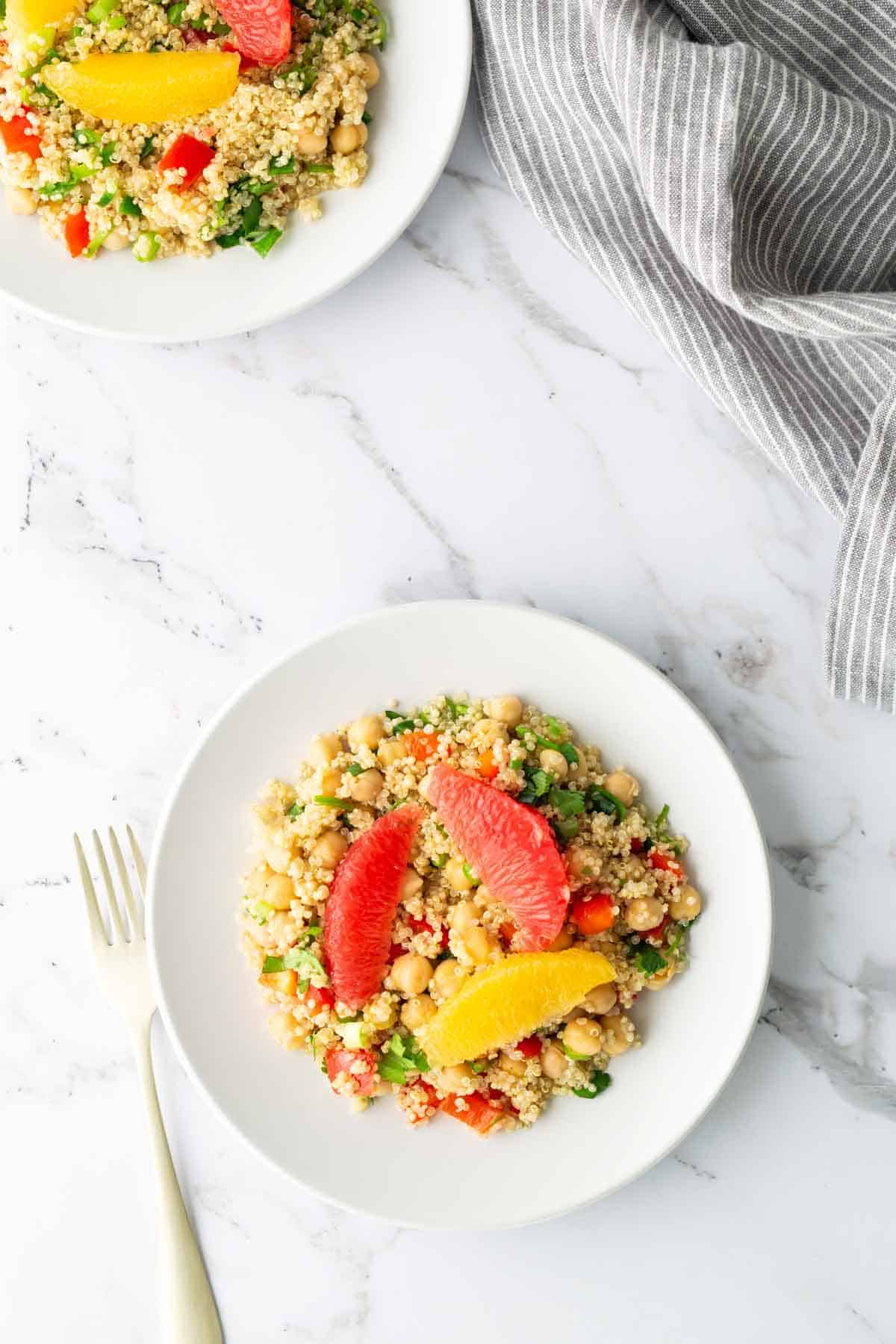 Two plates of chickpea and quinoa salad topped with grapefruit and orange slices, on a marble surface with a gray-striped cloth and a fork.