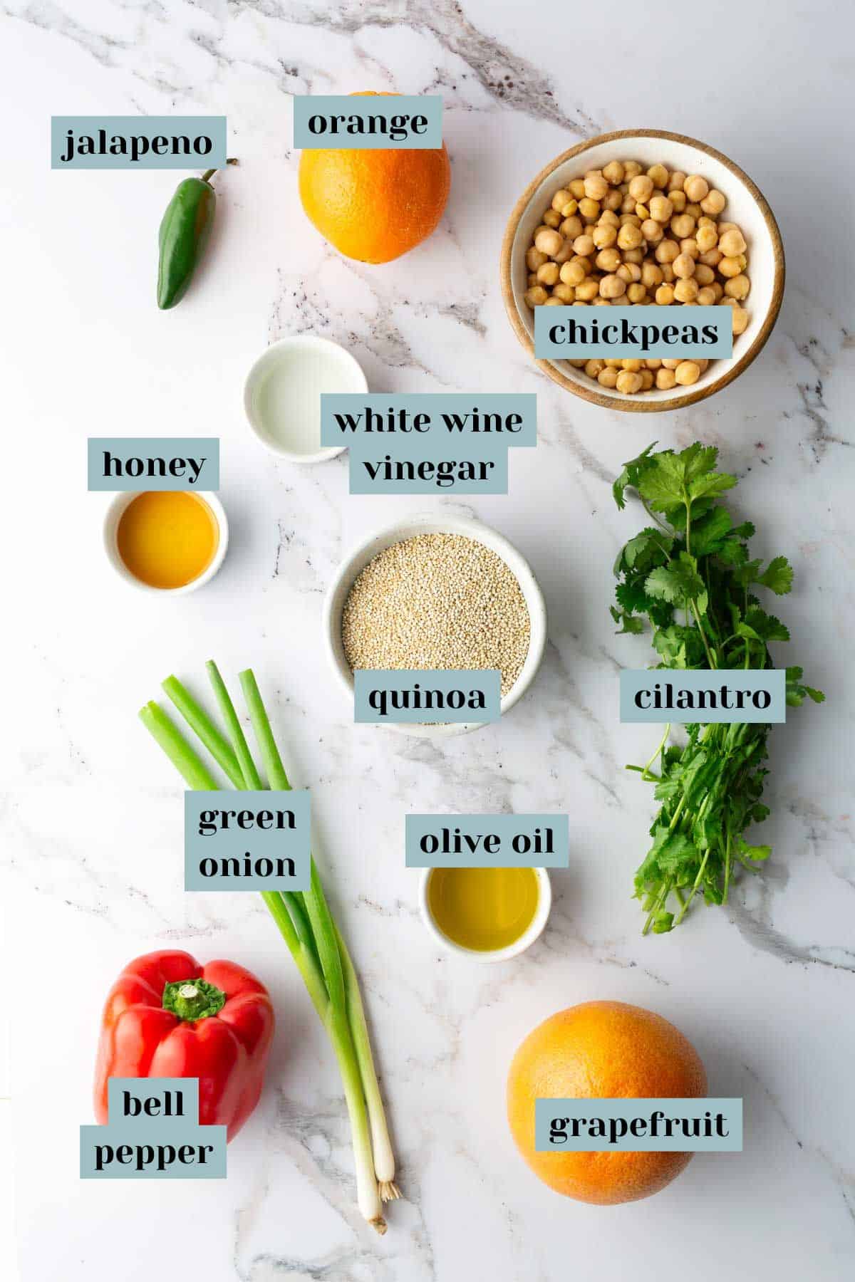 Ingredients laid out on a marble surface, including jalapeno, orange, chickpeas, honey, white wine vinegar, quinoa, cilantro, green onion, olive oil, bell pepper, and grapefruit.