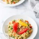 A white plate of quinoa salad featuring chickpeas, red and yellow bell peppers, garnished with orange and grapefruit slices. A fork is placed on the side. Another plate and a striped cloth are in view.