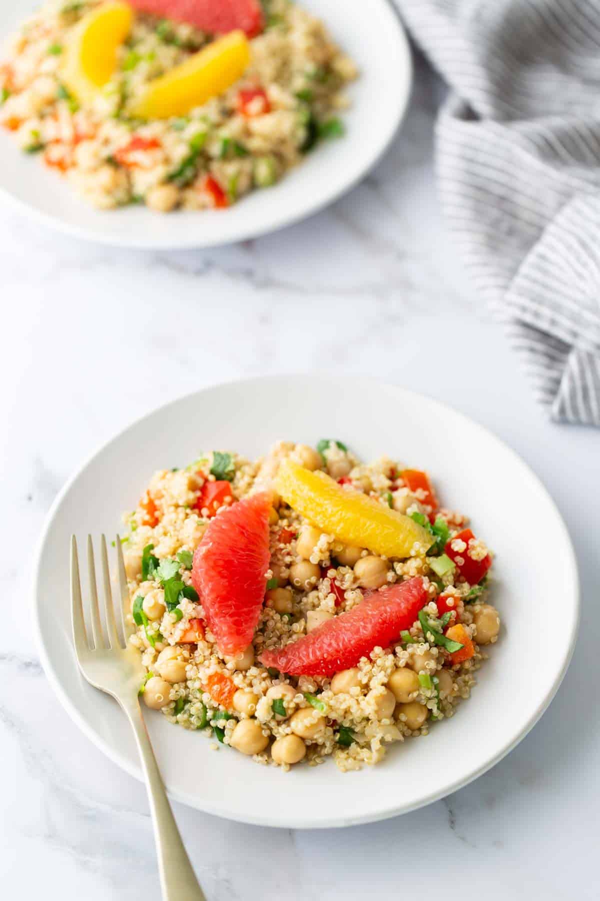 A white plate of quinoa salad featuring chickpeas, red and yellow bell peppers, garnished with orange and grapefruit slices. A fork is placed on the side. Another plate and a striped cloth are in view.