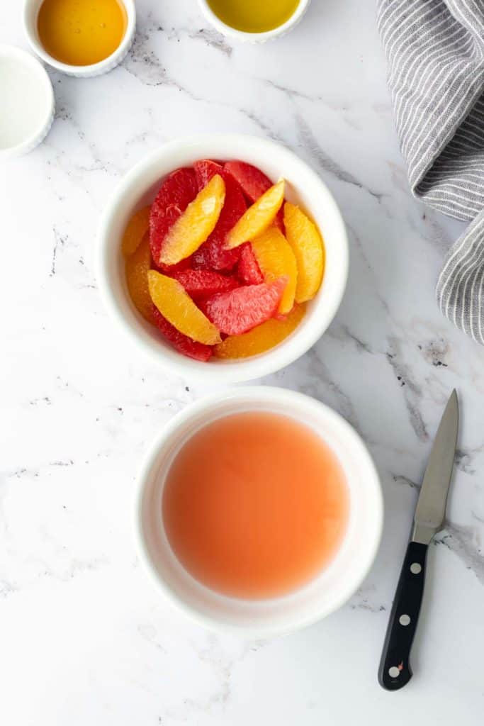 Two bowls on a marble countertop; one with sliced citrus fruits, the other containing juice. A small knife and a striped cloth are nearby.