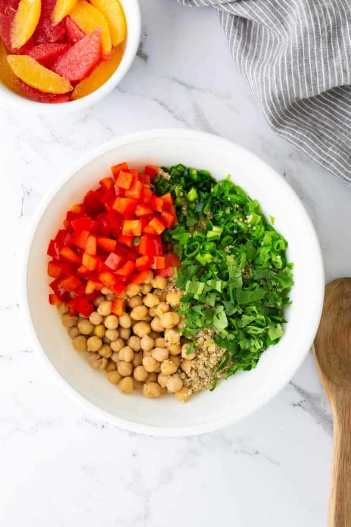 A white bowl with chickpeas, diced red bell pepper, chopped green onions, herbs, and seeds on a marble surface. A wooden spoon and a small bowl of citrus slices are nearby.