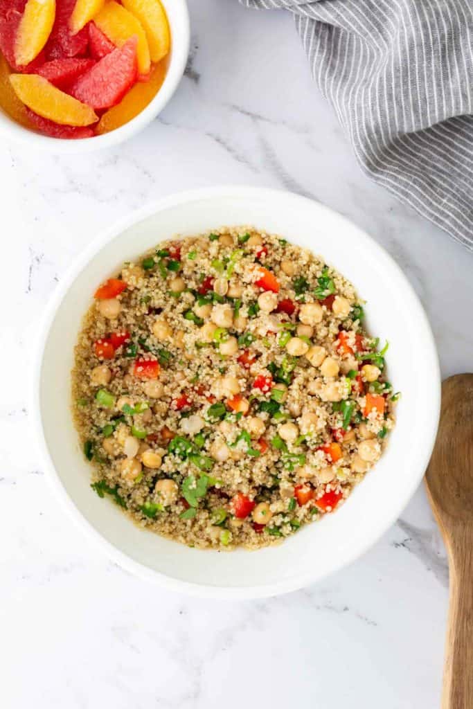 A bowl of quinoa salad with chickpeas, red bell peppers, and greens on a marble surface. A side dish of sliced citrus fruits is nearby.