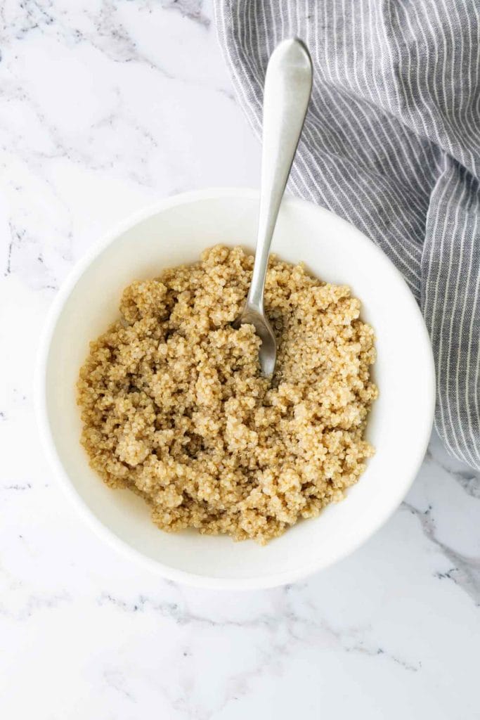 A bowl of cooked quinoa with a spoon on a marble surface, next to a striped cloth.