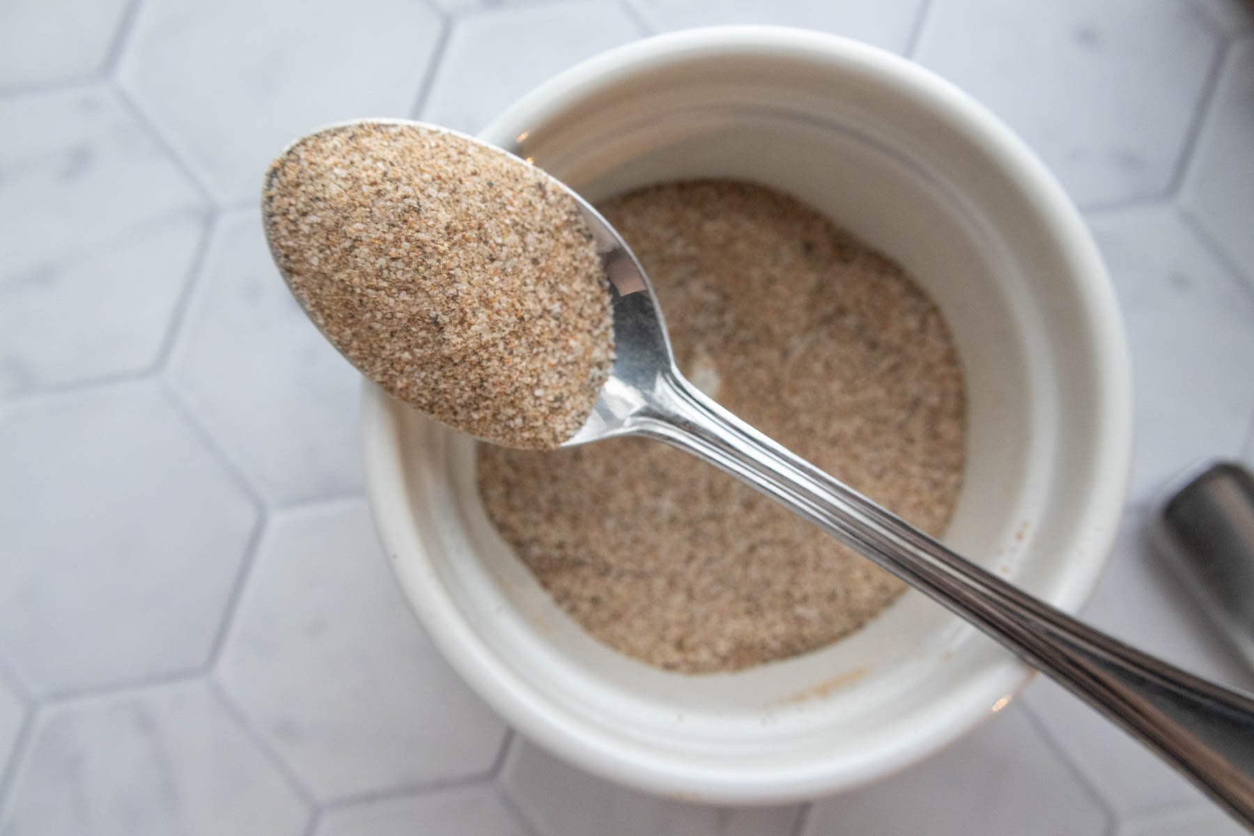 A spoonful of ground spices hovers above a white bowl containing more of the same spice mix. The background features a hexagonal tile surface.