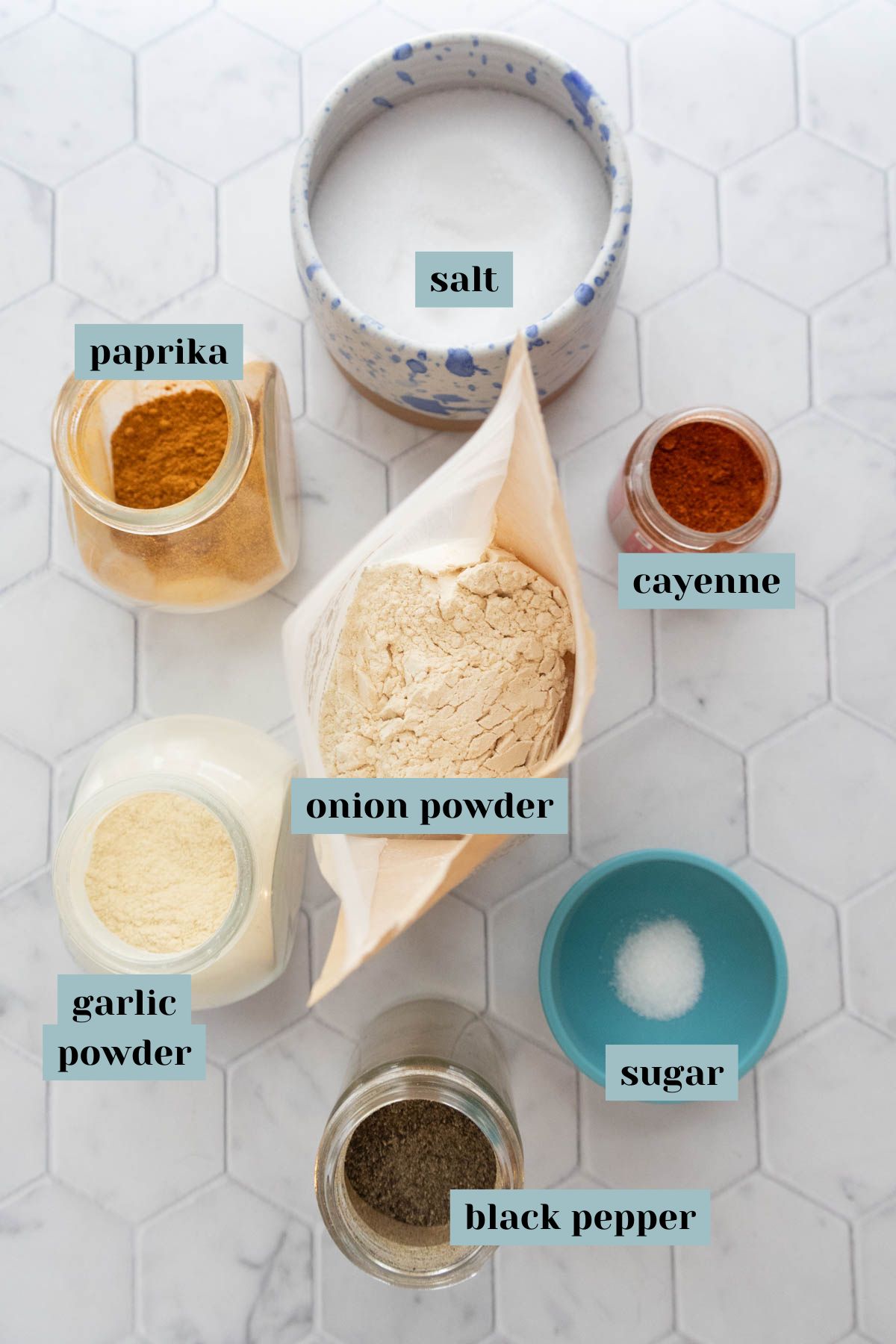 Assorted labeled spices on a hexagonal white tile surface, including salt, paprika, cayenne, onion powder, garlic powder, black pepper, and sugar.