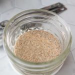 A small glass jar filled with a light brown, finely ground powder sits on a hexagonal tile surface.