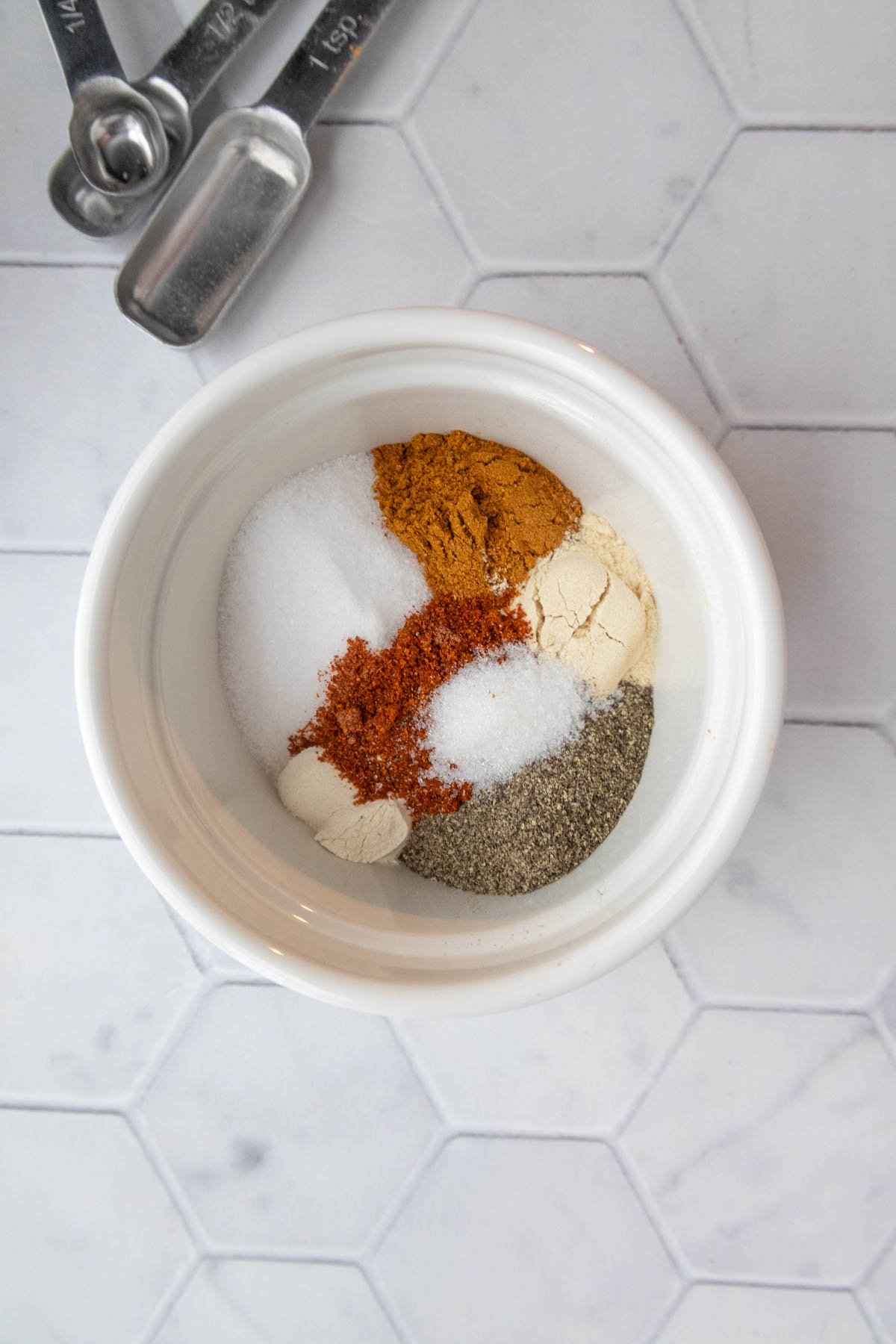 A white bowl with spices and salt is on a hexagon-tiled surface next to metal measuring spoons.