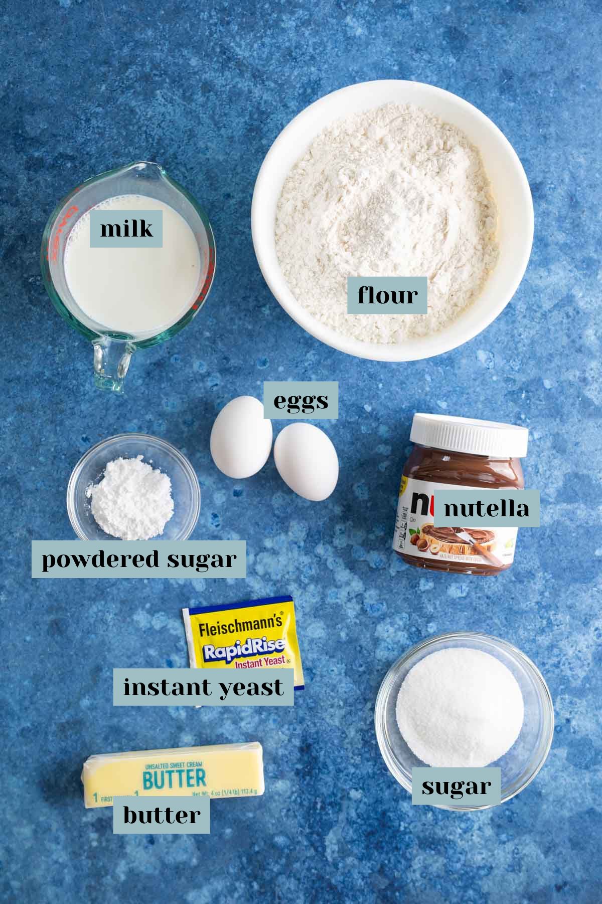 Ingredients for baking: flour, milk, eggs, Nutella, powdered sugar, instant yeast, butter, and sugar on a blue surface.