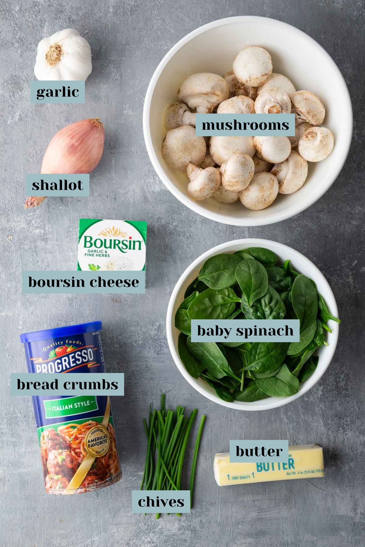 Ingredients for a recipe are arranged on a gray surface: garlic, shallot, mushrooms, boursin cheese, baby spinach, breadcrumbs, chives, and butter.