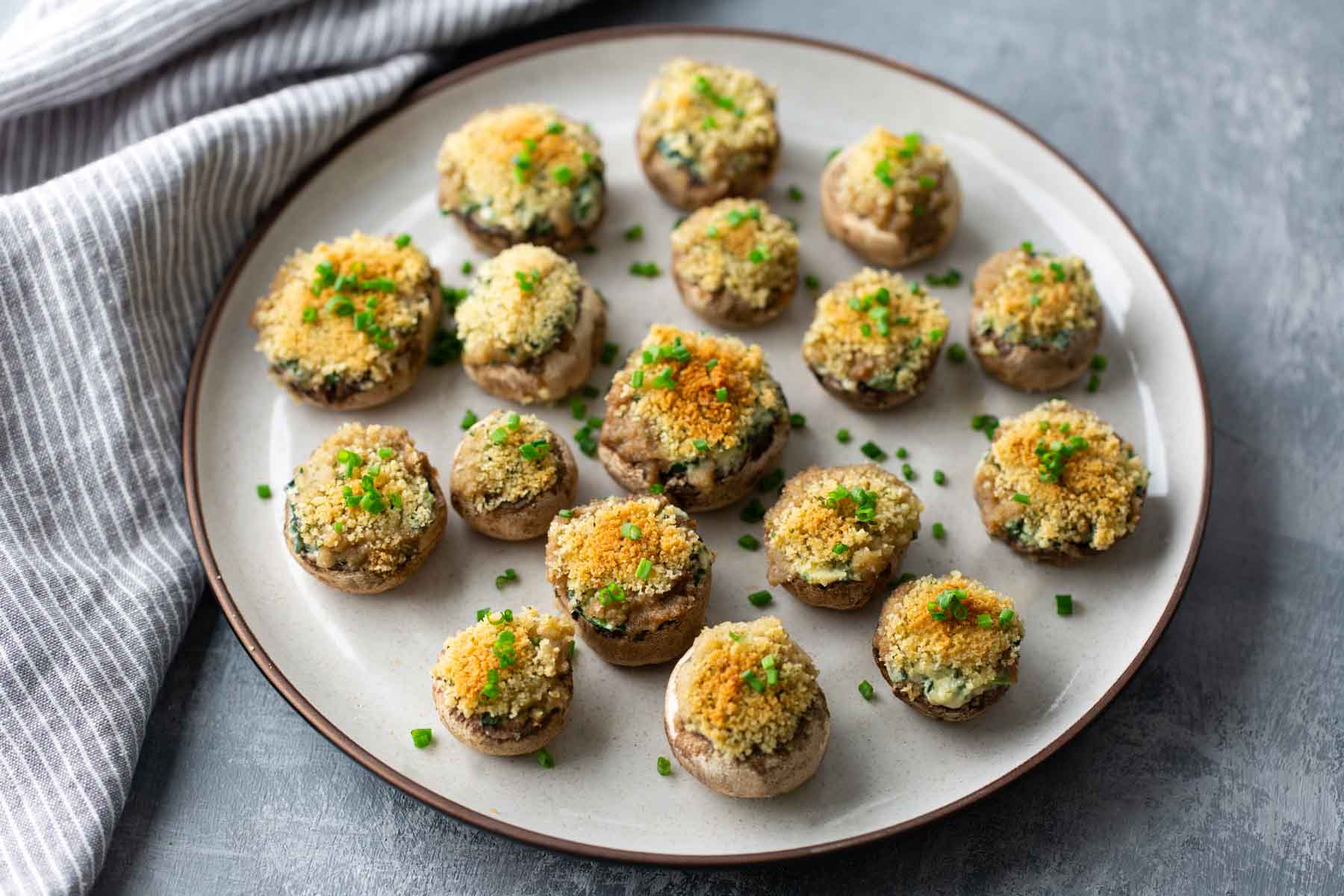 A plate of baked stuffed mushrooms topped with breadcrumbs and garnished with chopped chives, set on a gray tablecloth.