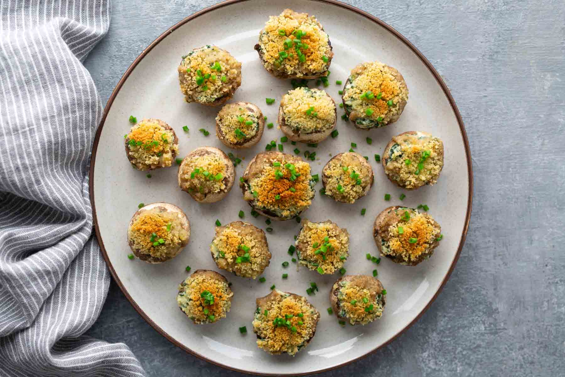 A plate of stuffed mushrooms topped with breadcrumbs and chopped herbs, arranged in rows on a gray surface. A striped cloth is partially visible on the side.