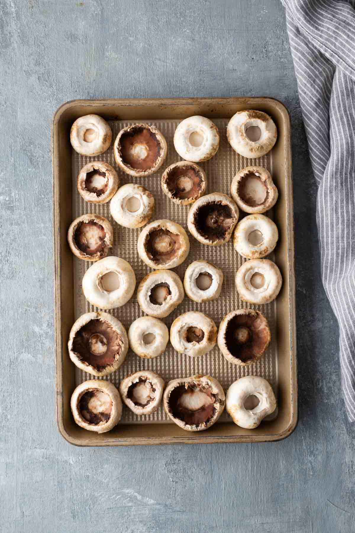 A baking tray filled with evenly spaced, uncooked mushroom caps, arranged in rows on a textured gray surface. A striped cloth is visible on the right side.