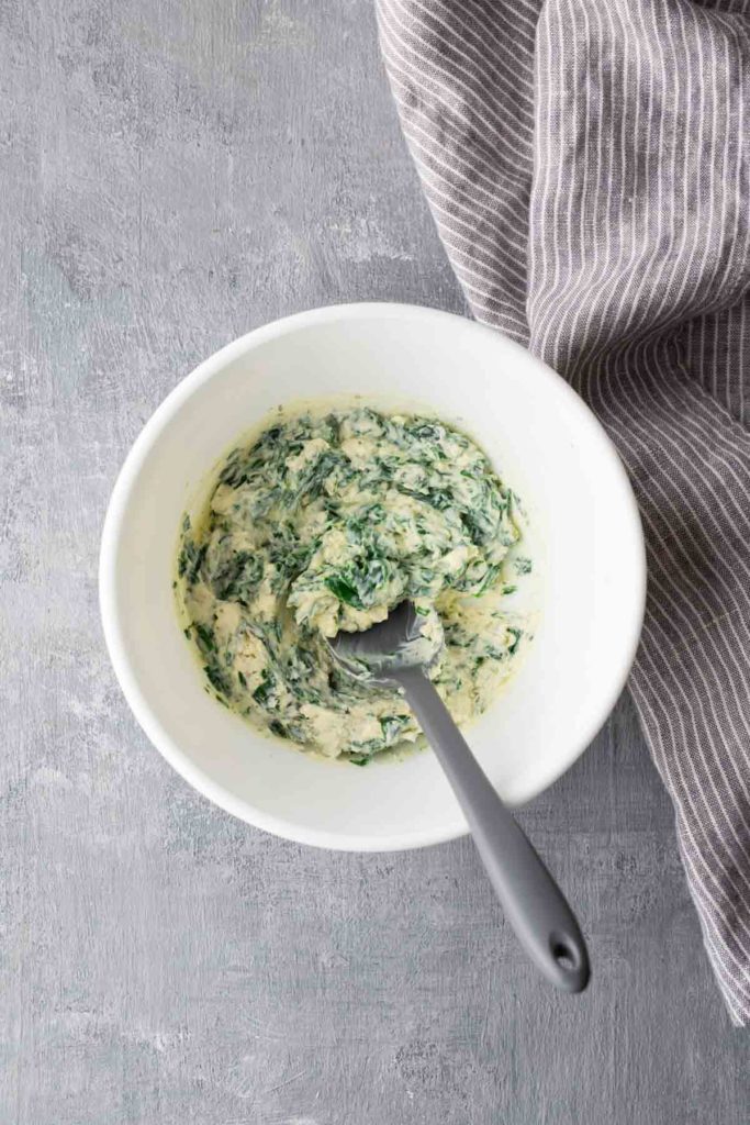 A white bowl with spinach and cheese mixture, placed on a gray surface. A spoon rests inside the bowl. A striped cloth is on the right side.