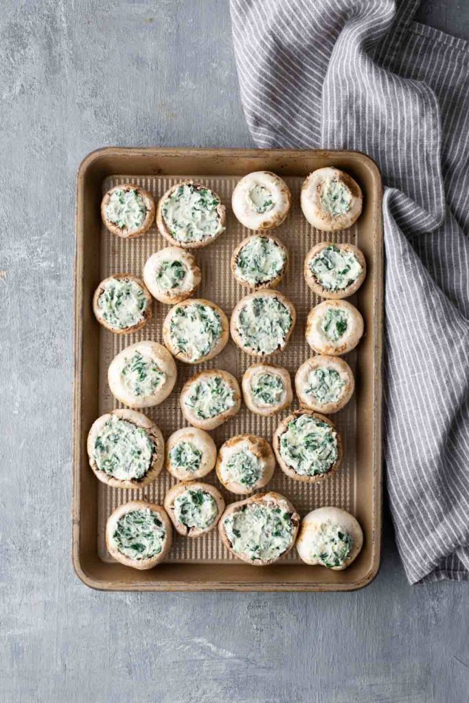 A baking tray filled with stuffed mushrooms, topped with a green and white filling, next to a gray striped cloth on a textured surface.