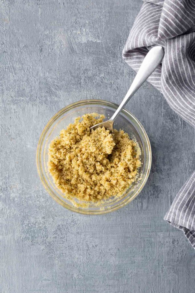 A bowl of cooked quinoa with a spoon in it is placed on a textured surface beside a striped cloth.