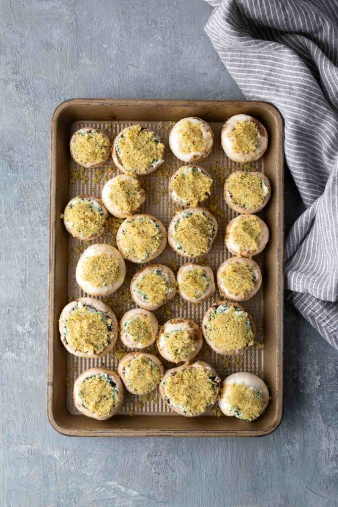 A baking tray filled with uncooked stuffed mushrooms topped with breadcrumbs, placed on a textured surface with a striped cloth beside it.