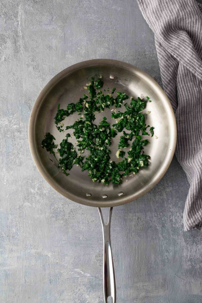 Chopped spinach sautéing in a stainless steel pan on a gray surface, with a striped cloth nearby.