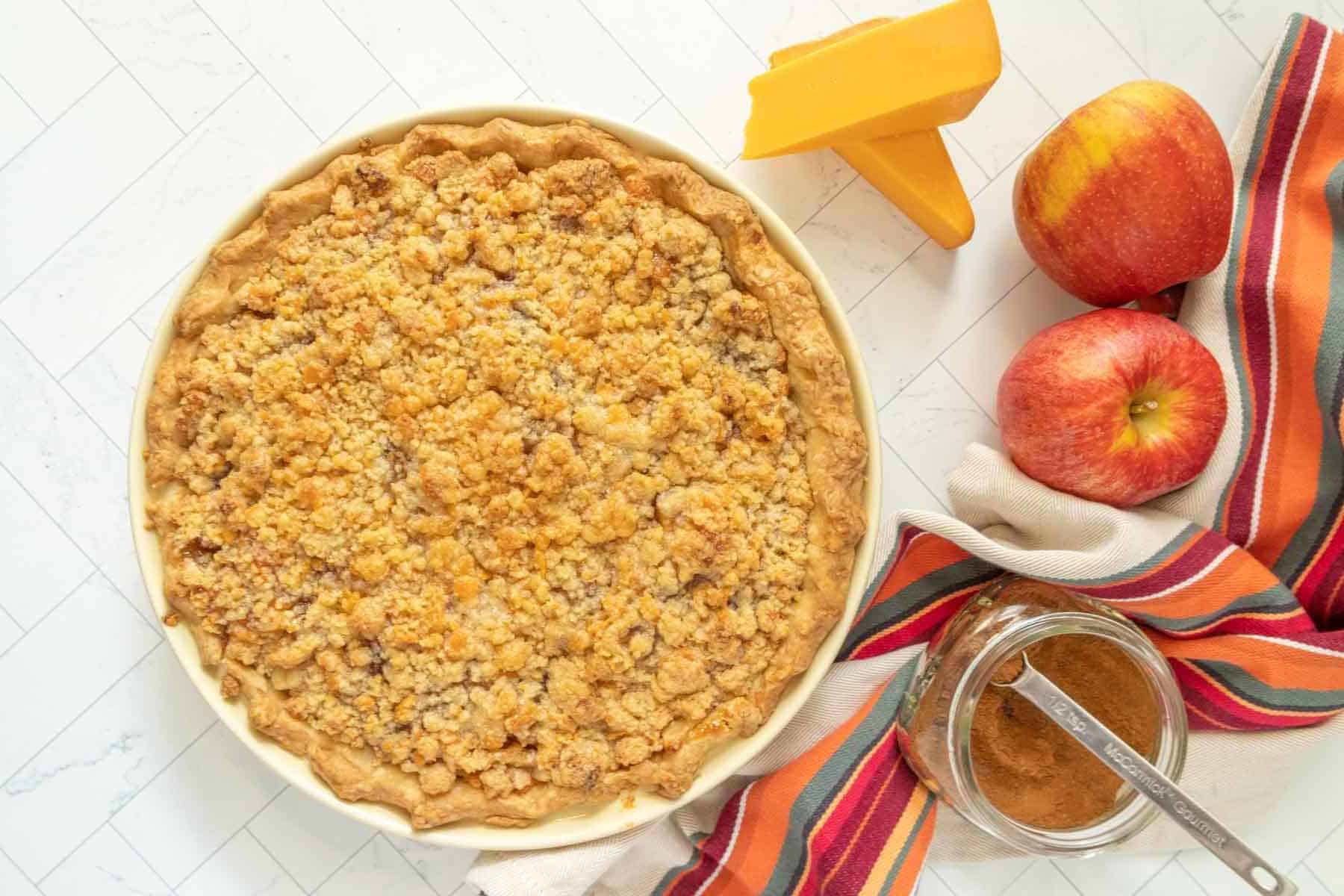 A pie is sitting on a table next to apples and cheese.