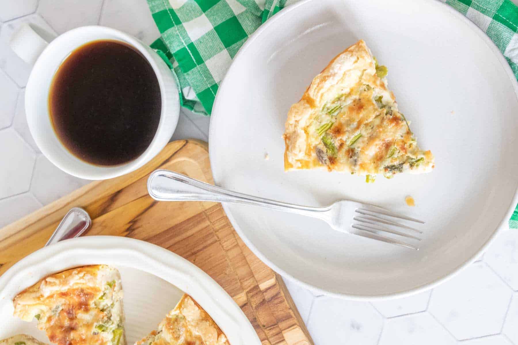 Overhead photo of a slice of asparagus quiche on a gray plate with a cup of coffee beside.