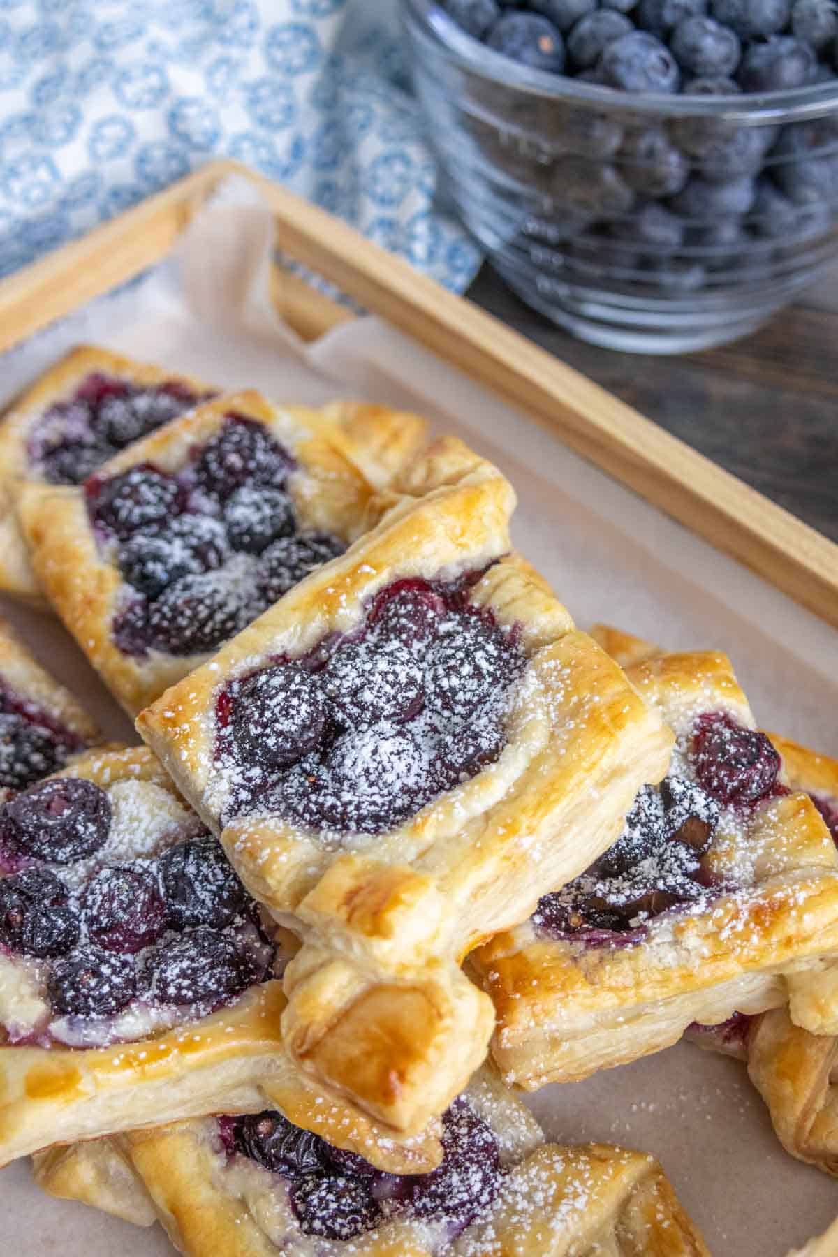 Rectangular blueberry pastries dusted with powdered sugar are arranged on a wooden tray lined with parchment paper. A bowl of fresh blueberries is in the background.