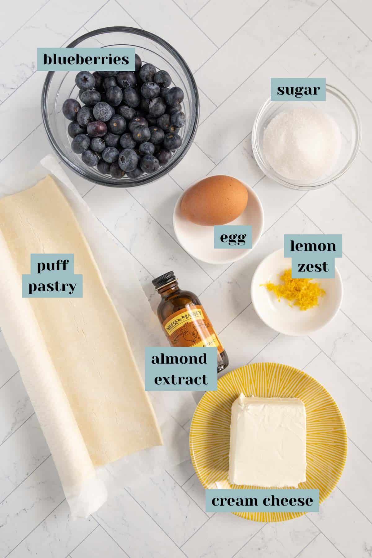 Ingredients for a dessert on a white surface: blueberries, sugar, egg, lemon zest, puff pastry, almond extract, and cream cheese.