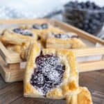 Close-up of a puff pastry with powdered sugar and blueberry filling. More pastries and a bowl of blueberries are blurred in the background, all on a wooden surface.