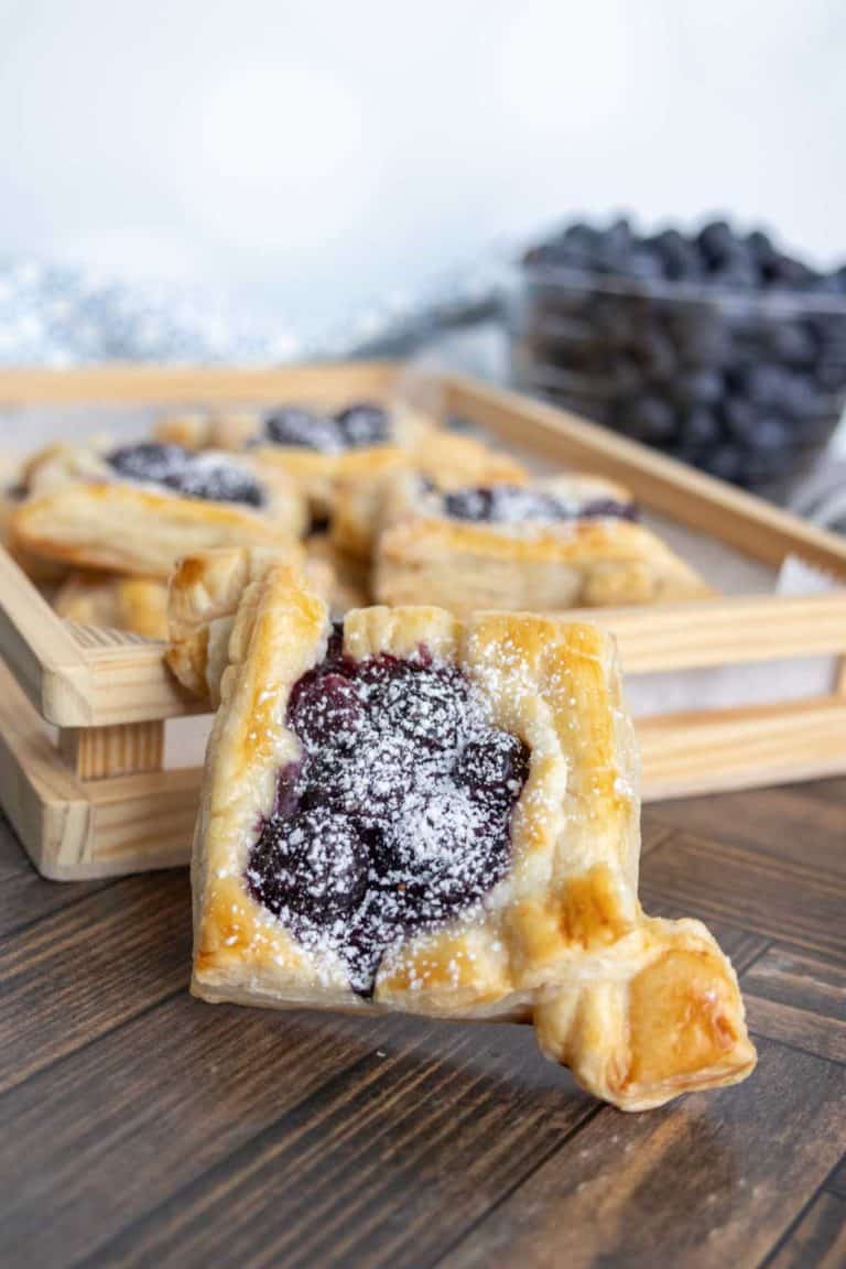 Close-up of a puff pastry with powdered sugar and blueberry filling. More pastries and a bowl of blueberries are blurred in the background, all on a wooden surface.