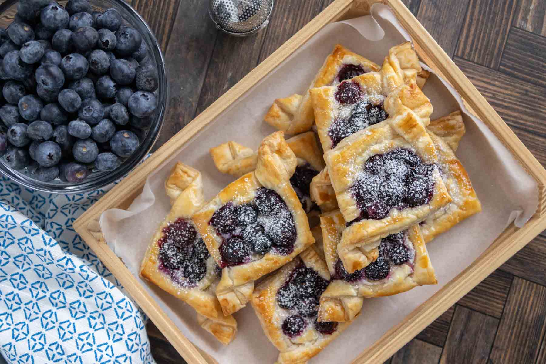 A tray of blueberry pastries dusted with powdered sugar next to a bowl of fresh blueberries on a wooden table.