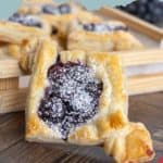 Blueberry Danish on a wooden surface, garnished with powdered sugar. Other Danishes in the background.