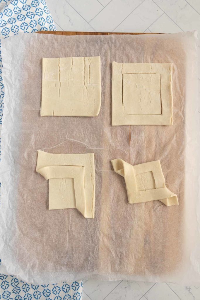 Four pieces of pastry dough on parchment paper, two with rectangular cuts, one partially folded, on a light-colored surface.