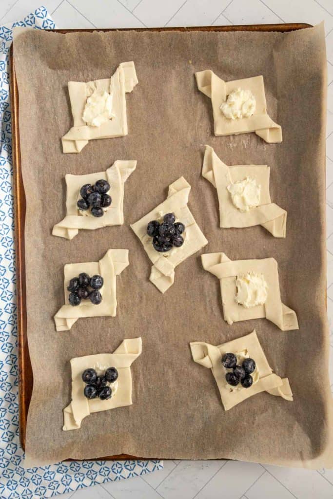 Unbaked pastries on a baking tray lined with parchment paper, topped with ricotta and blueberries.