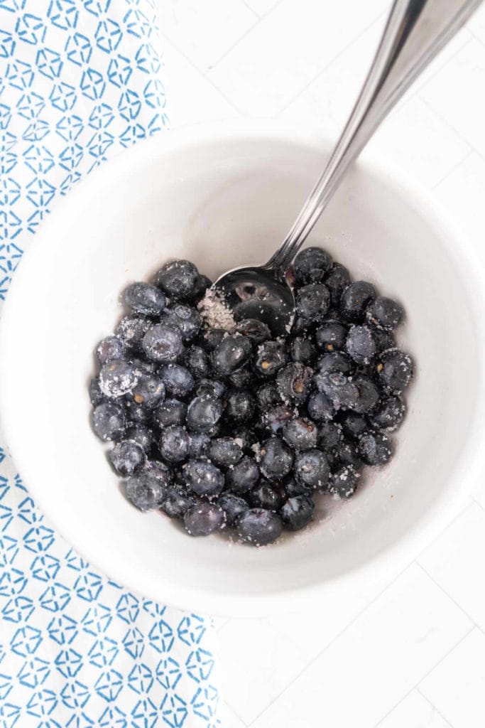 White bowl filled with sugared blueberries and a metal spoon, placed on a patterned blue and white surface.