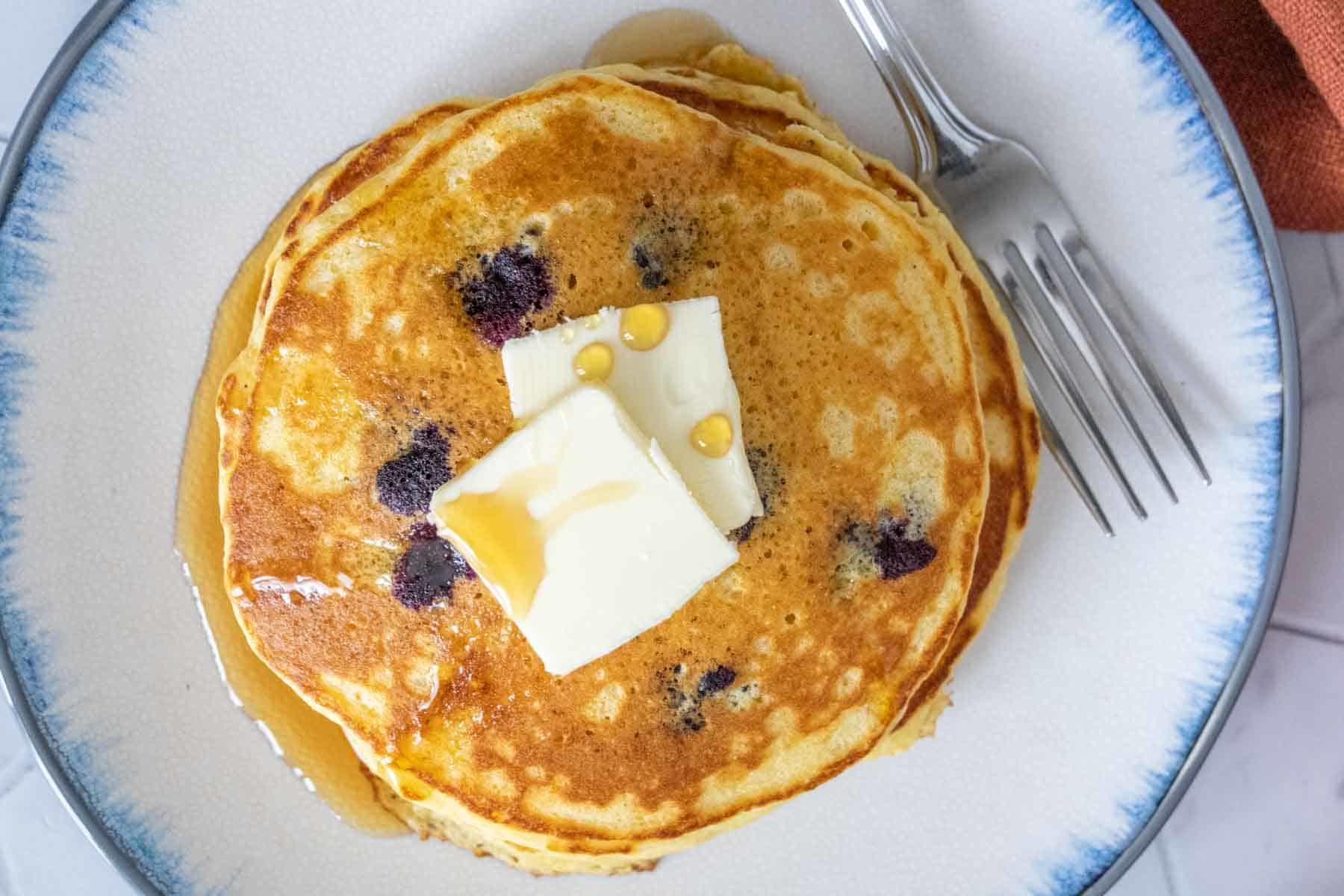 Overhead of plate of blueberry pancakes with butter and syrup on top with a fork beside.