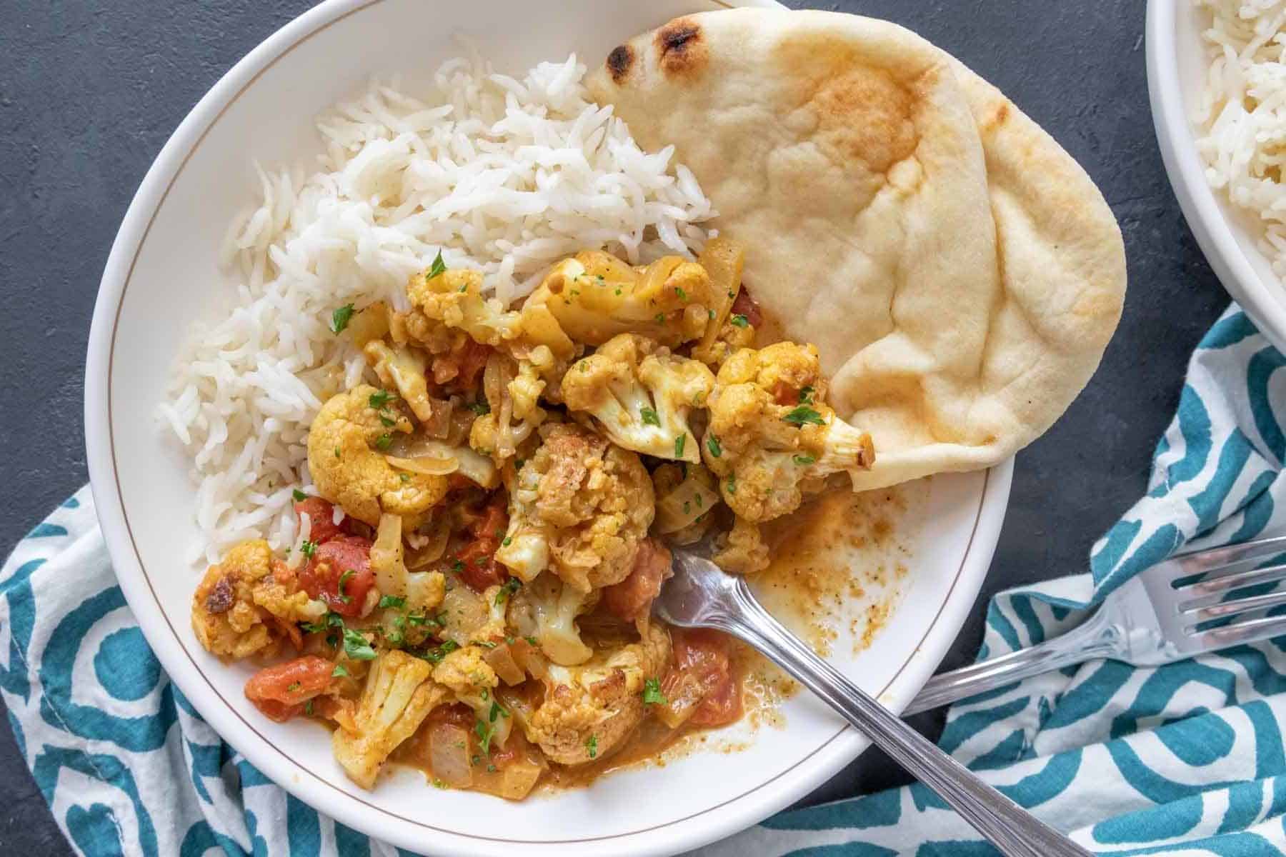 Overhead photo of cauliflower curry in a white bowl with rice and naan.