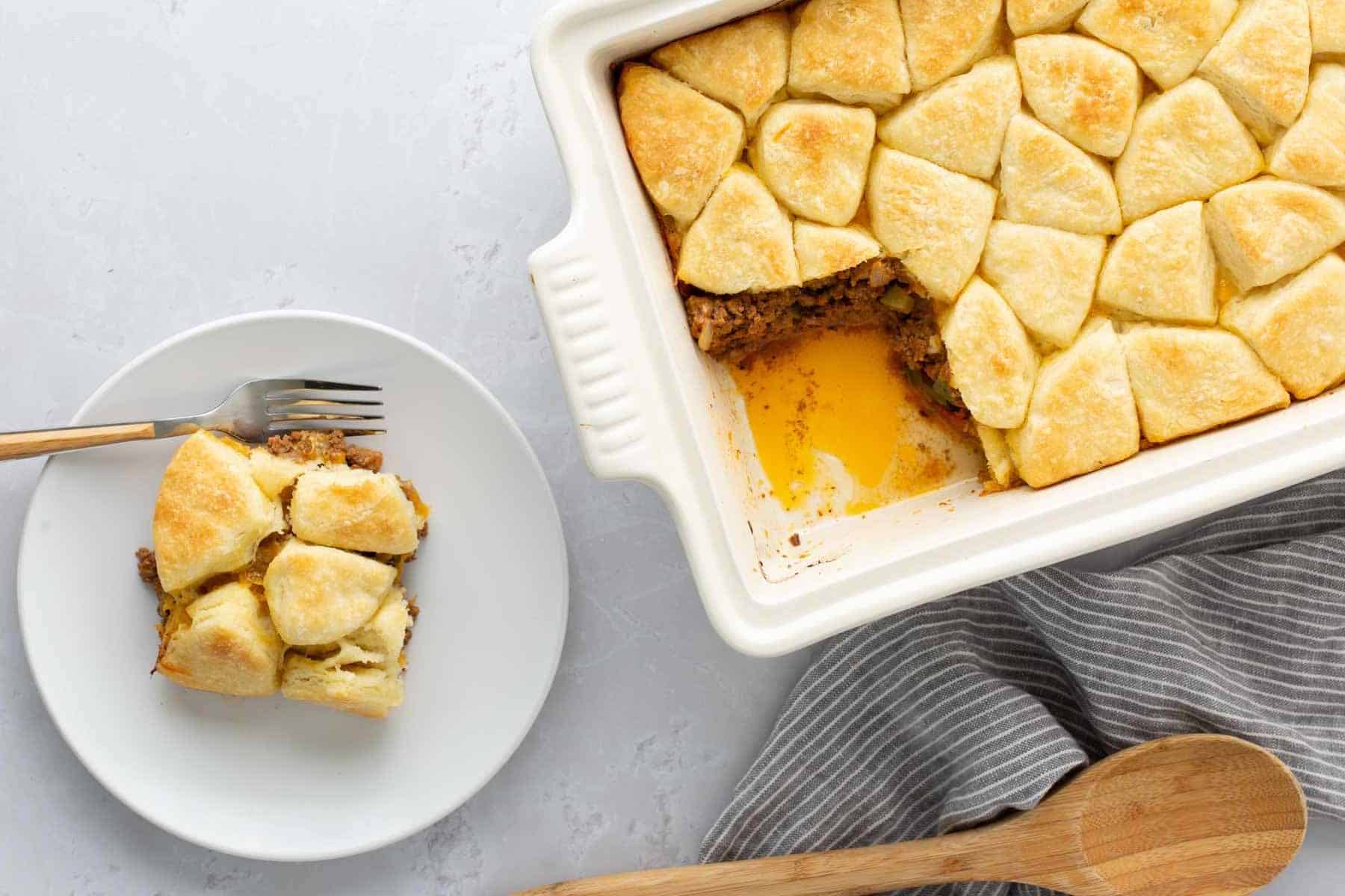 A casserole dish filled with a baked, golden brown biscuit-topped casserole. One serving is placed on a white plate with a fork. A striped cloth and wooden spoon are beside the dish.