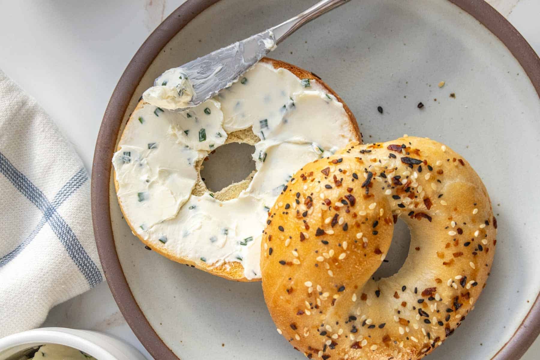 An everything bagel with cream cheese on a plate, half of it spread with cream cheese, beside a knife with cream cheese.