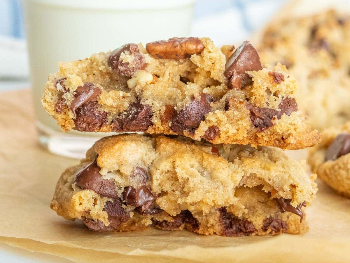 A glass of milk is placed behind two stacked chocolate chip cookies on a piece of parchment paper.