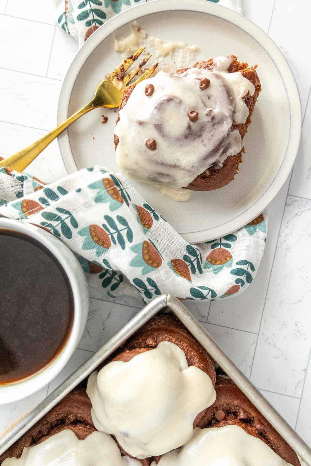 A cinnamon roll with icing and chocolate chips on a plate next to a fork, a cup of coffee, and a tray of more rolls. A patterned cloth is draped nearby.