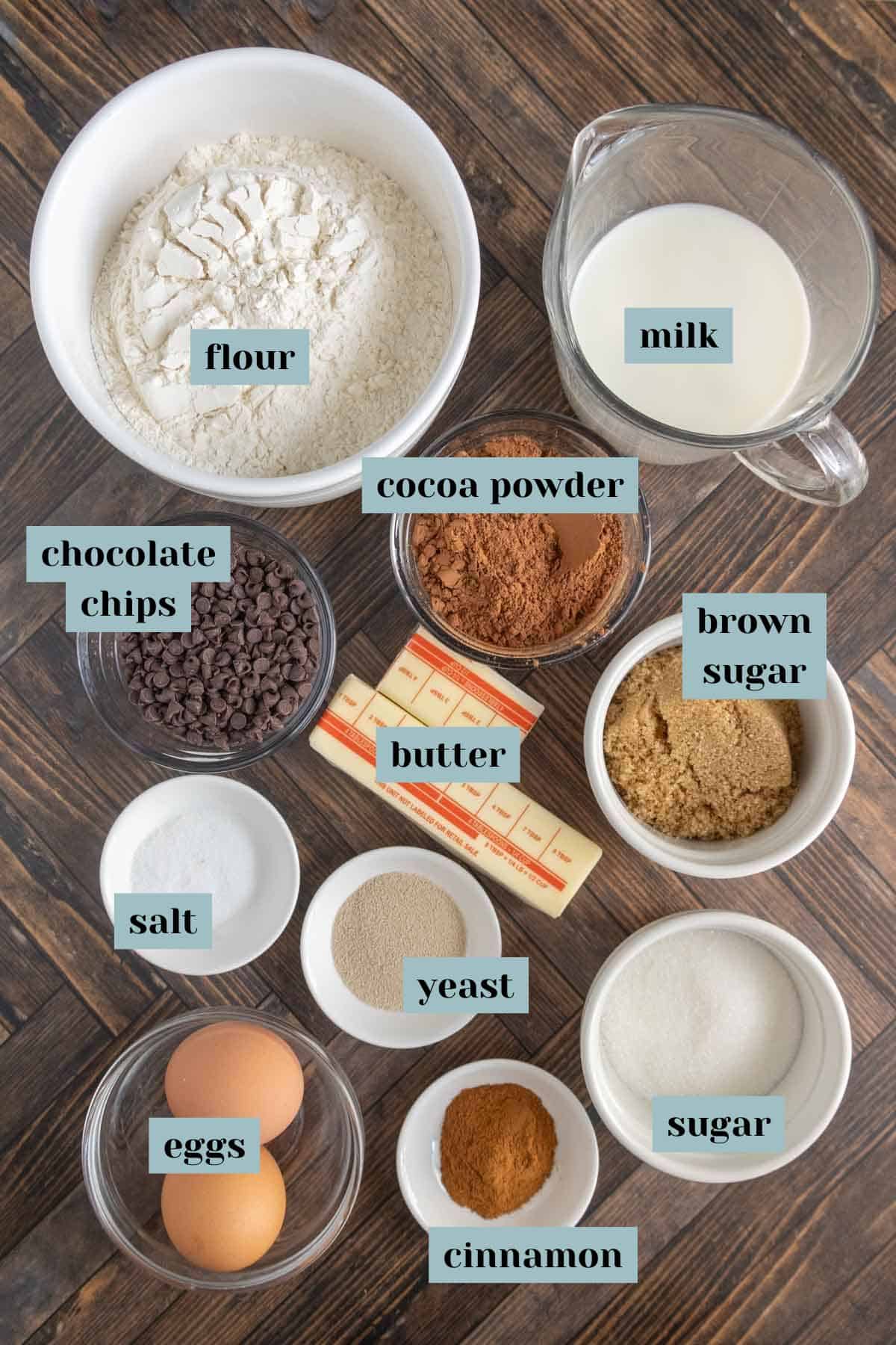 Assorted baking ingredients including flour, milk, cocoa powder, brown sugar, butter, chocolate chips, salt, yeast, eggs, cinnamon, and sugar arranged on a wooden surface.