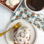 Cinnamon roll with icing on a plate next to a cup of coffee. More cinnamon rolls in a tray nearby. A gold fork rests on the plate over a patterned napkin.