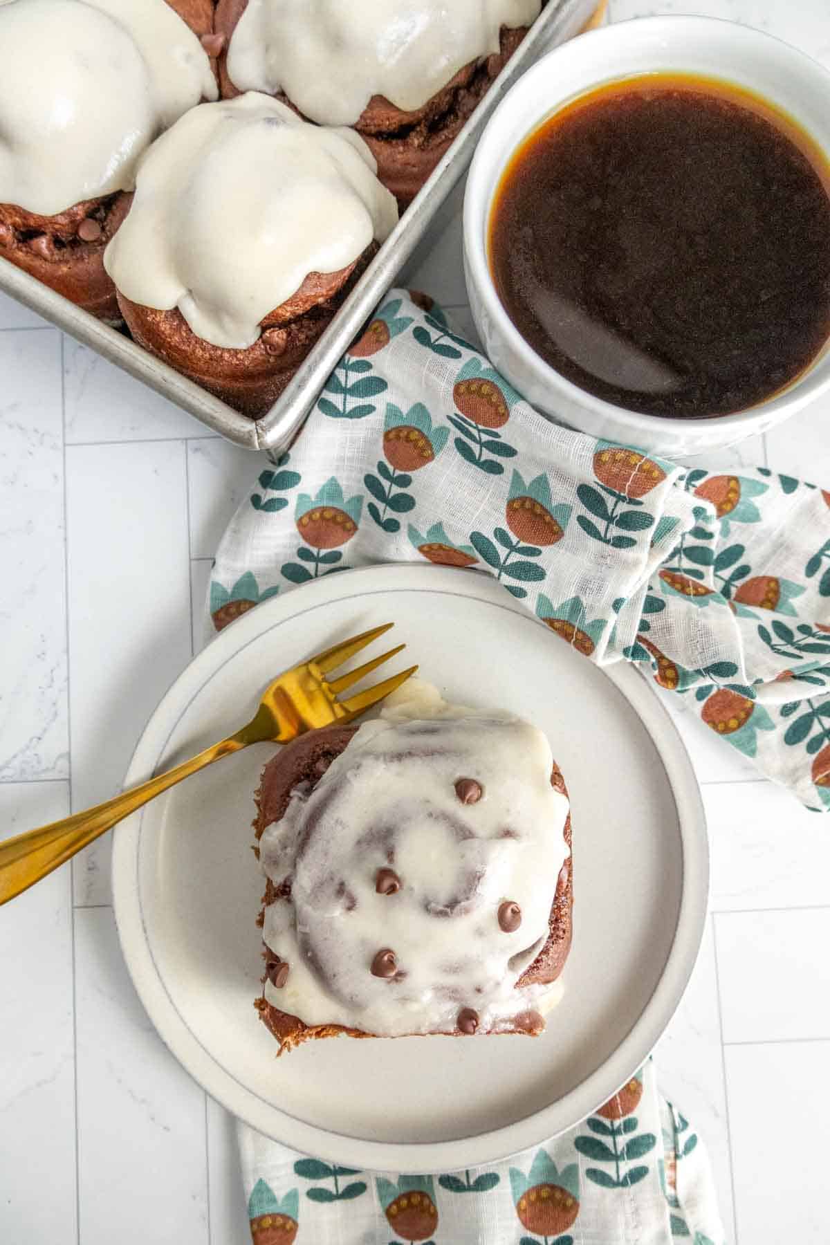 Cinnamon roll with icing on a plate next to a cup of coffee. More cinnamon rolls in a tray nearby. A gold fork rests on the plate over a patterned napkin.