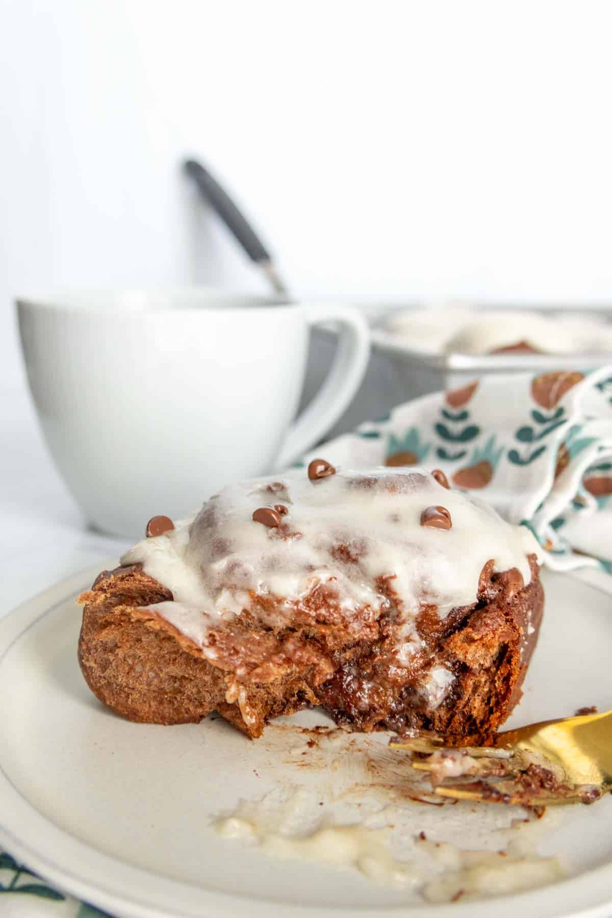 Cinnamon roll with white icing on a plate, partially eaten, with a gold fork beside it. In the background, a white mug and a patterned cloth are visible.