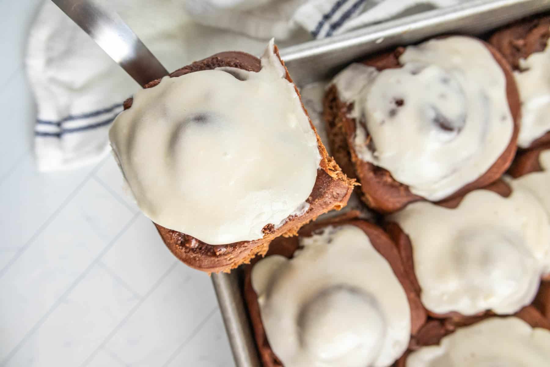 Close-up of a tray of frosted cinnamon rolls, with one roll being lifted by a spatula.