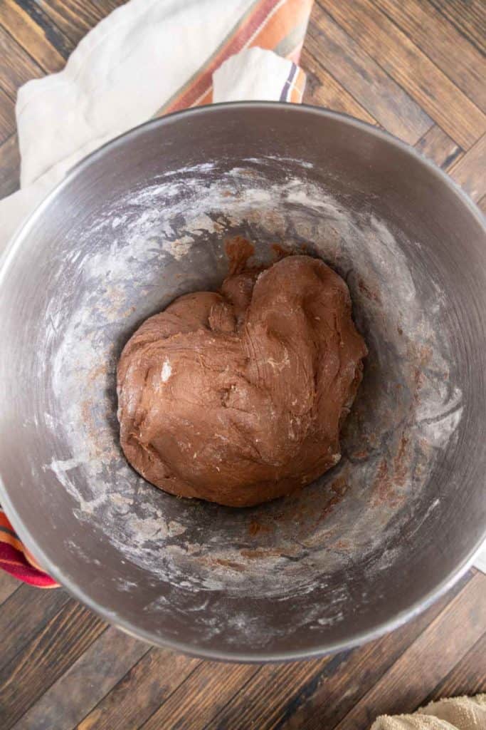 A bowl with chocolate cookie dough on a wooden surface, next to a folded cloth.