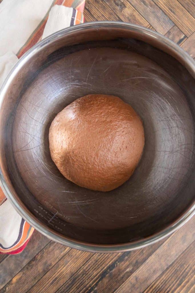 Ball of chocolate dough in a metal mixing bowl on a wooden surface with a cloth nearby.