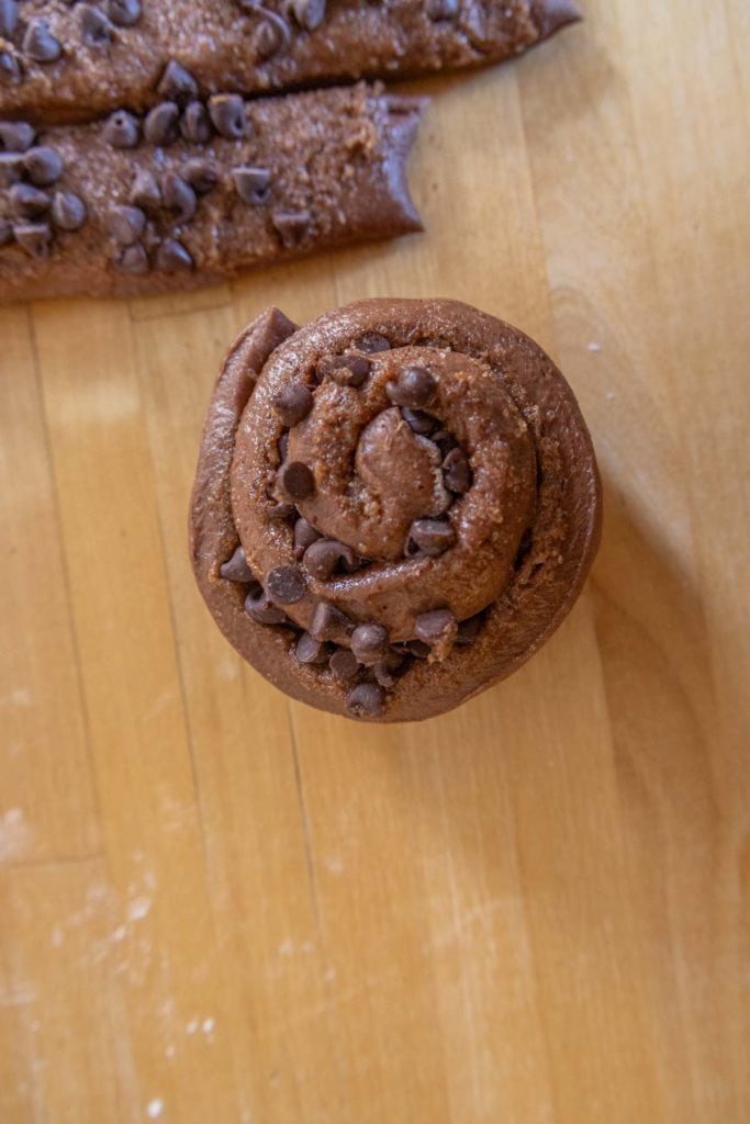 A cinnamon roll with chocolate chips on a wooden surface. In the background, dough strips with chocolate chips are visible.