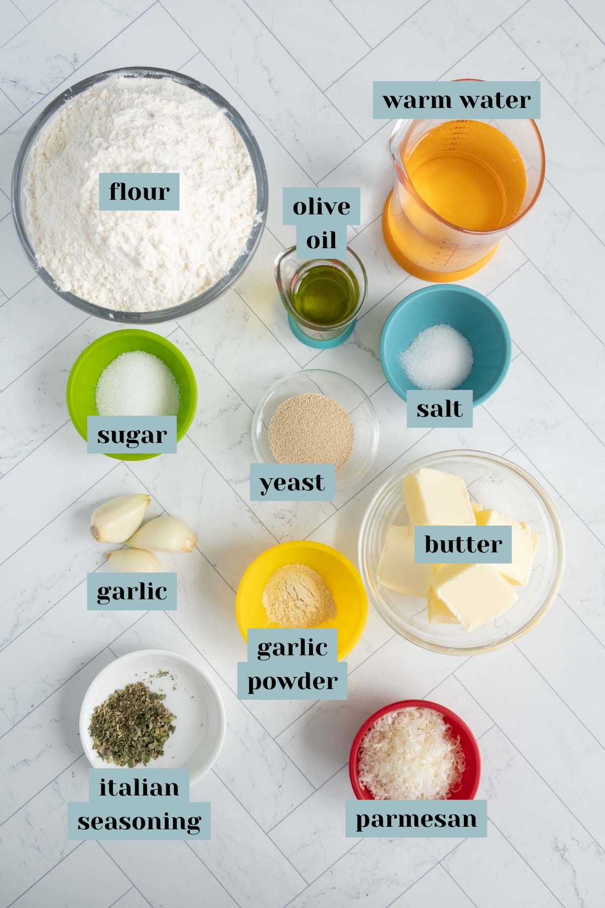 Top-down view of baking ingredients labeled in bowls: flour, olive oil, warm water, sugar, salt, yeast, butter, garlic, garlic powder, Italian seasoning, parmesan.