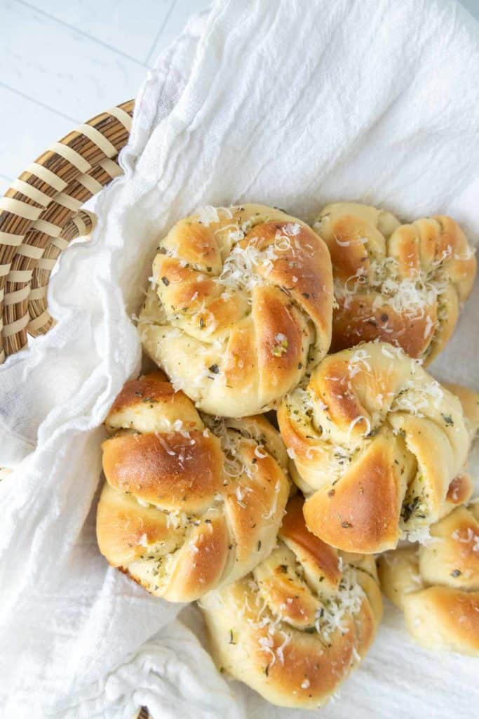 A basket of freshly baked garlic knots topped with grated cheese, resting on a white cloth.